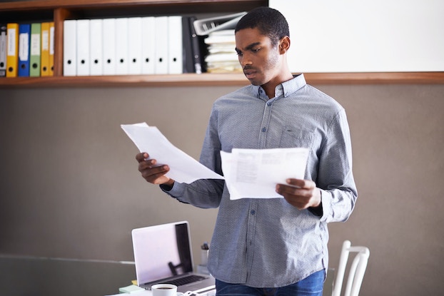 An entrepreneur is standing in an office and holding multiple sheets of paper in both of his hands. He has an expression of concern.